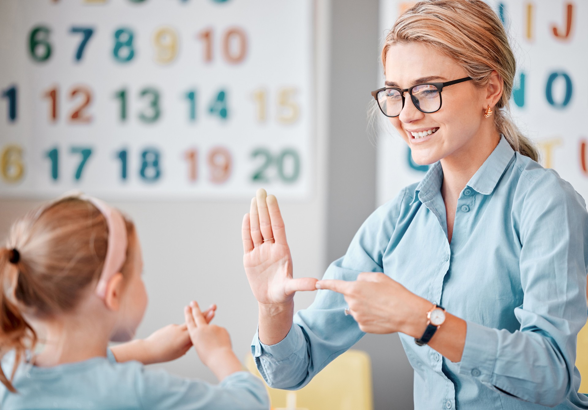 Smiling caucasian teacher wearing spectacles communicating with deaf girl student with hand sign language in a classroom at school. Sign language teacher in a tutoring class for a cute female child with hearing disability